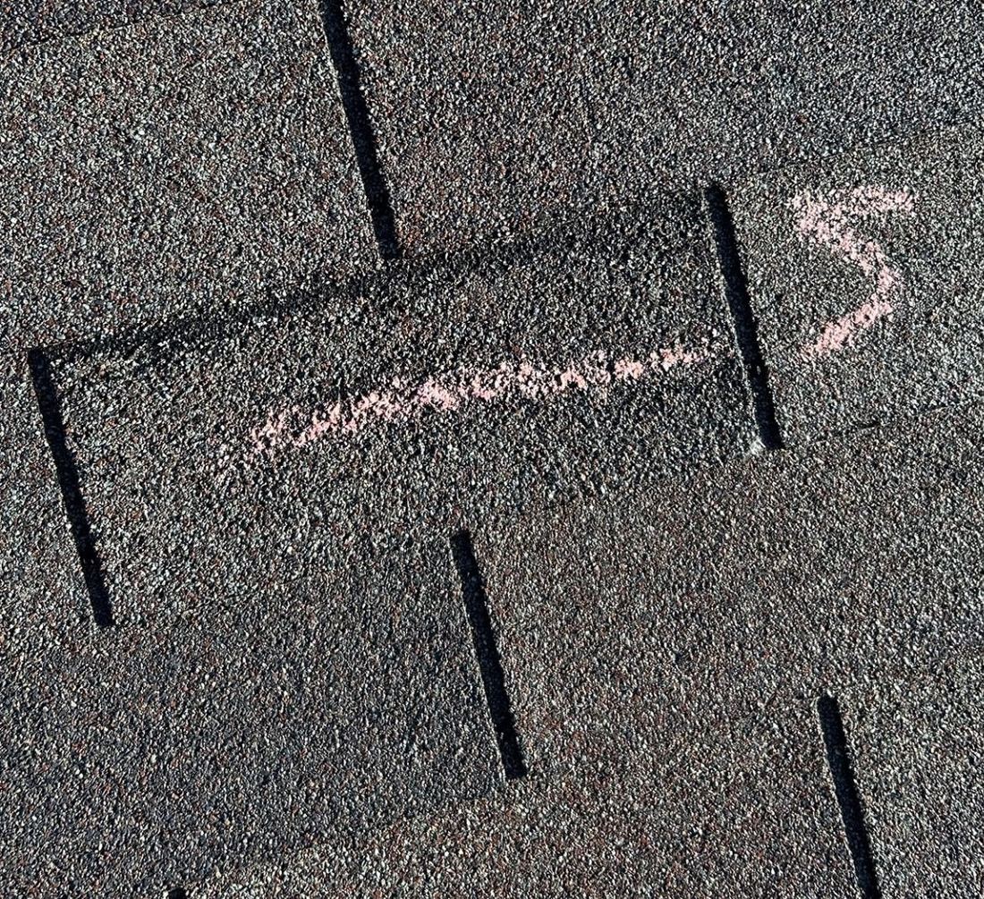 A 3-tab shingle tab with a diagonal dark scuff band marked with chalk, one course below the ridge vent